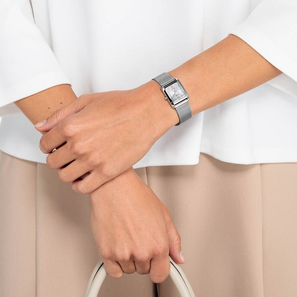 Frau mit silberner Uhr, weißer Handtasche, beiger Hose und weißem Hemd.
